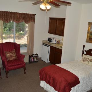 A cozy bedroom featuring a bed, a comfortable chair, and a kitchenette area, illuminated by natural light from the window.