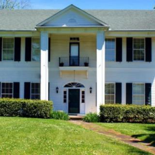 A two-story colonial-style house with white exterior and black shutters, surrounded by a green lawn.