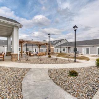 A peaceful courtyard with a central pathway, outdoor seating, and surrounding buildings under a cloudy sky.