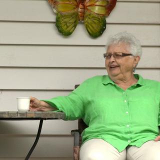 An elderly woman in a green shirt sits outside with a coffee cup, smiling next to a butterfly decoration.
