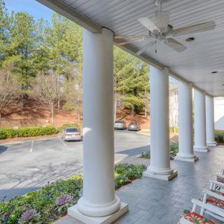 A porch with large columns overlooking a driveway and parked cars, surrounded by trees.