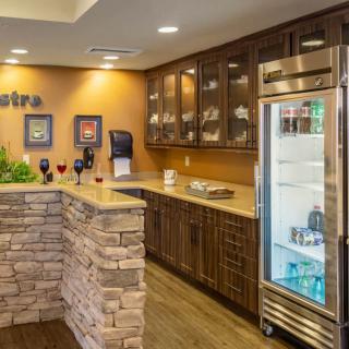 A modern kitchen area with a stone-designed counter and wooden cabinets, featuring wine glasses and a glass-front refrigerator.