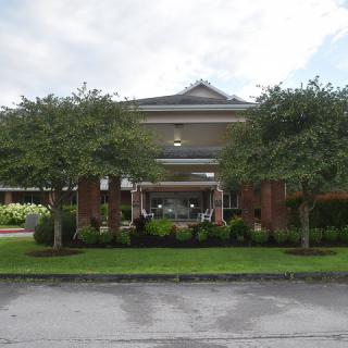 A brick building with a covered entrance, surrounded by landscaped greenery and trees under a partly cloudy sky.