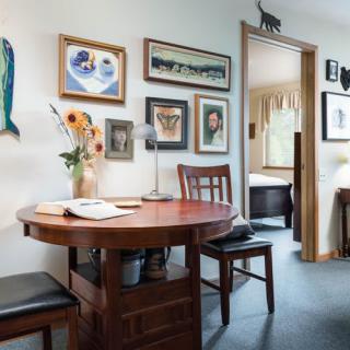 A cozy interior space featuring a round dining table with chairs, surrounded by various artworks and decorations on the wall. A vase of sunflowers adds a touch of freshness to the room.