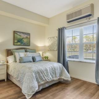 A cozy bedroom with a neatly made bed, curtains drawn back from a window and an air conditioner above.