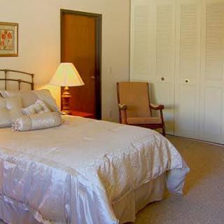A cozy bedroom featuring a neatly made bed with pillows, a lamp on a side table, and an armchair beside the closet.