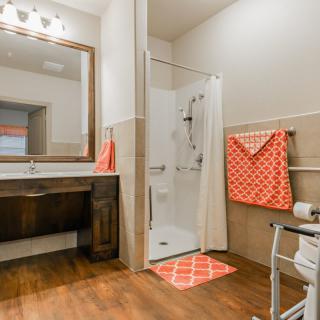 A modern accessible bathroom with a walk-in shower, wooden flooring, and orange towels.