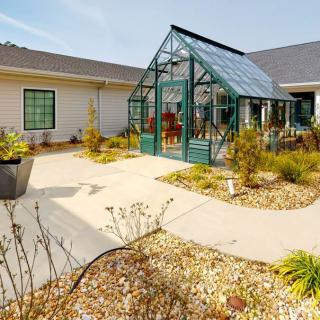 A small greenhouse in a garden setting, surrounded by various plants and pathways.