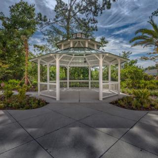 A white gazebo surrounded by lush greenery and trees under a partly cloudy sky.