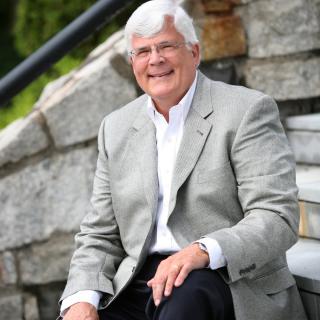 A man with grey hair, dressed in a suit, is sitting on outdoor steps and smiling.
