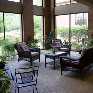 A cozy lounge area featuring wicker furniture, surrounded by large windows and lush indoor plants.