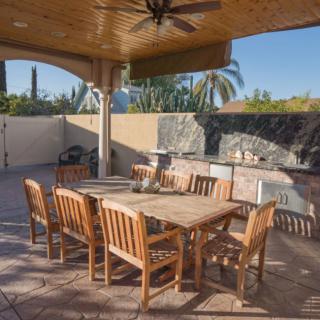 A spacious outdoor patio with a wooden dining set under a covered terrace, featuring a ceiling fan and surrounded by palm trees.