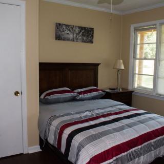 A cozy bedroom featuring a neatly made bed with striped bedding, a nightstand with a lamp, and a window allowing natural light to filter in.
