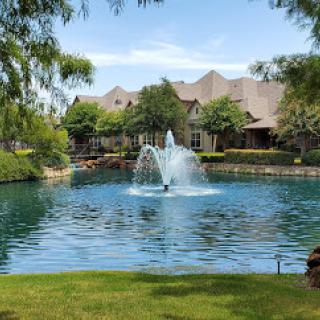 A serene view of a residential lake featuring a beautiful fountain, surrounded by lush greenery and clear blue skies.