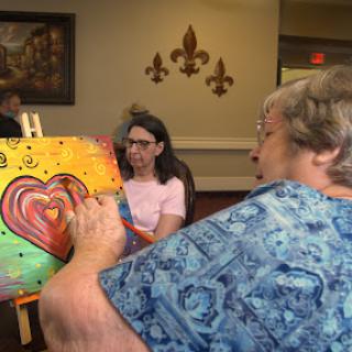An elderly woman focuses on painting a colorful heart on her canvas while others engage in a creative art session.
