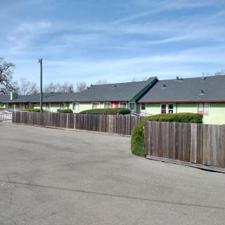 A row of motel buildings with a wooden fence alongside a wide road under a clear blue sky.