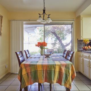 A cozy dining area with a colorful tablecloth and flowers on the table, well-lit by natural sunlight through large windows.