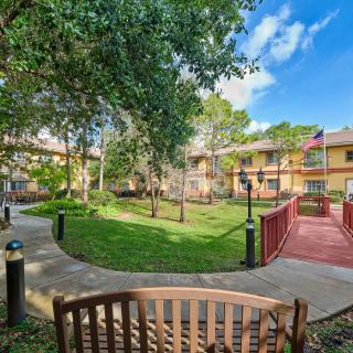 A tranquil garden with a wooden bench, surrounded by trees and buildings under a clear blue sky.