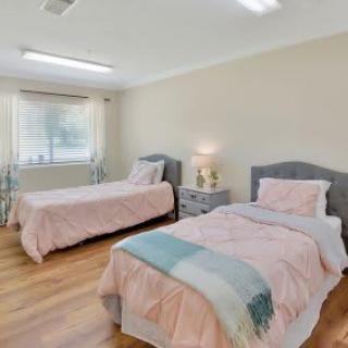 A cozy bedroom featuring two twin beds with pink bedding, a nightstand with a lamp, and a window allowing natural light.