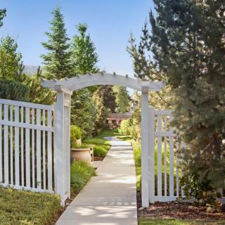 A scenic garden pathway with a white arch and fence, surrounded by lush greenery.