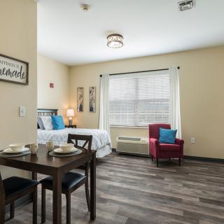 A modern bedroom with a bed, dining table, and armchair, featuring large windows and stylish decor.