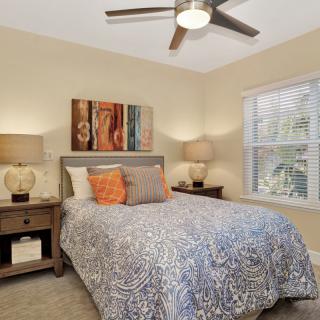 A cozy bedroom features a neatly arranged bed with patterned bedding, flanked by two lamps on nightstands. Artwork adorns the wall above the headboard, and natural light filters through a window with blinds.