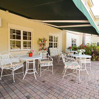 A cozy outdoor patio with metal tables and chairs beneath a green awning, surrounded by lush plants.