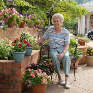 An elderly woman sitting on a patio surrounded by colorful potted flowers and plants.