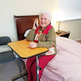 A smiling senior woman sits by a bed, talking on the phone with a mobile table in front of her.