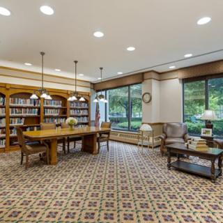 A cozy library reading room with bookshelves, a central wooden table, and large windows.