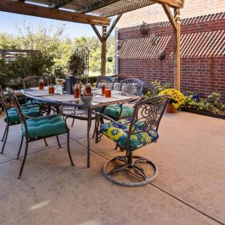 A cozy outdoor patio setup with a table and chairs under a pergola, surrounded by plants and a brick wall.