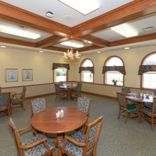 A well-lit dining room featuring round wooden tables, cushioned chairs, large windows, and a central chandelier.