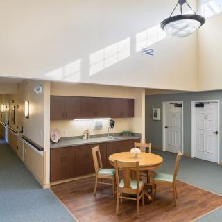 A minimalist interior featuring a dining area with a round table and four chairs, adjacent to a kitchenette and hallway.