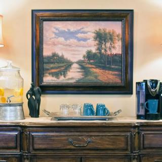 A cozy interior featuring a landscape painting above a wooden sideboard with a water dispenser, glasses, and a coffee maker.
