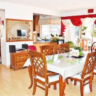 A cozy dining room with wooden furniture and a view into the kitchen, adorned with red curtains and indoor plants.