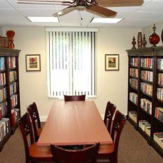 A cozy reading room featuring bookshelves filled with books, a large table surrounded by chairs, and ample natural light from the window.