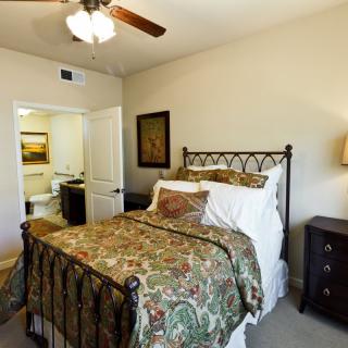 A warmly decorated bedroom featuring a double bed with patterned bedding, a bedside lamp, and an open doorway leading to another room.
