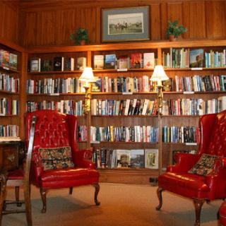A cozy library room with red armchairs, surrounded by wooden bookshelves filled with books and lit by warm lamps.