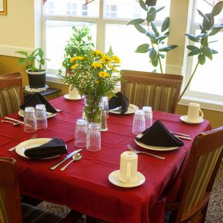 A neatly set dining table with flowers as a centerpiece and black napkins on a red tablecloth.