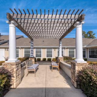 A serene outdoor patio featuring a pergola with white columns, complemented by seating and a clear blue sky.