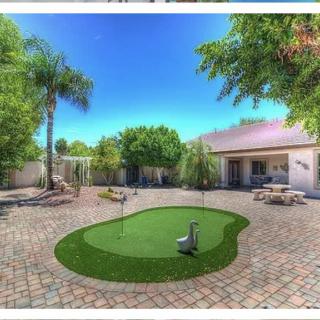 A spacious backyard with a putting green, surrounded by trees and outdoor seating on a paved patio.