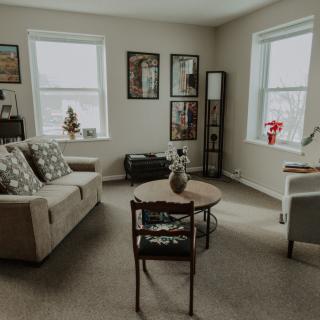 A cozy living room with a sofa, coffee table, and wall art, illuminated by natural light from two windows.