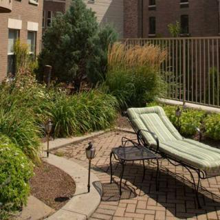 A rooftop garden with a lounge chair surrounded by lush greenery and a brick patio.