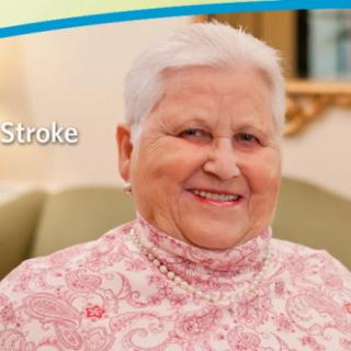 An elderly woman smiling indoors, wearing a pink blouse.