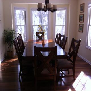 A dining room featuring a wooden table with chairs, illuminated by natural light from large windows.