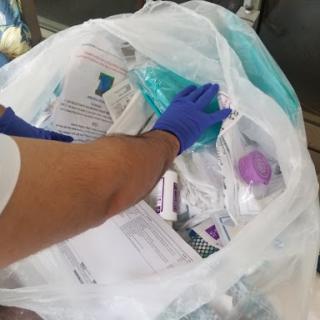 A gloved hand sifting through a bag filled with medical waste and disposable items.