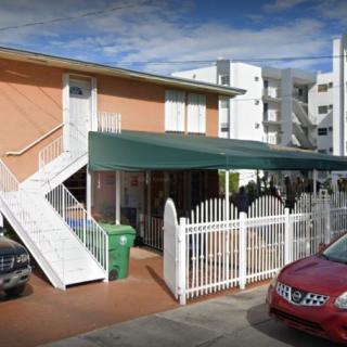 A two-story residential building with an external staircase and a green awning covering the entrance area, surrounded by cars and a white fence.