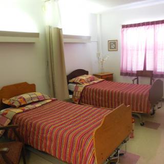 A hospital room featuring two twin beds with striped bedding and a window adorned with curtains.