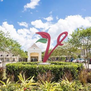 A vibrant sculpture stands in front of a shopping center, surrounded by lush greenery under a clear blue sky.