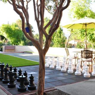 An outdoor patio with a giant chess set, surrounded by lush garden greenery and furniture under a sunny sky.